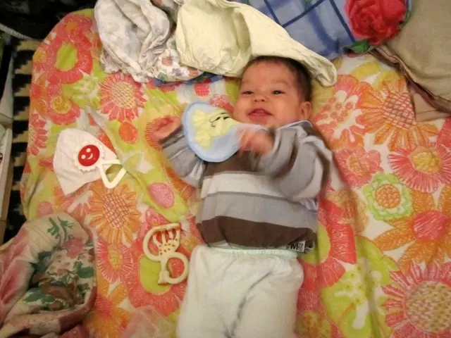 In this image there is a baby lying on the bed. Beside him there are toys, pillows and a few...