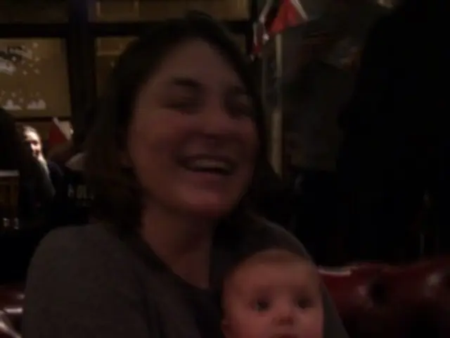 In this picture there is a woman sitting and smiling and she is holding the baby. At the back there...