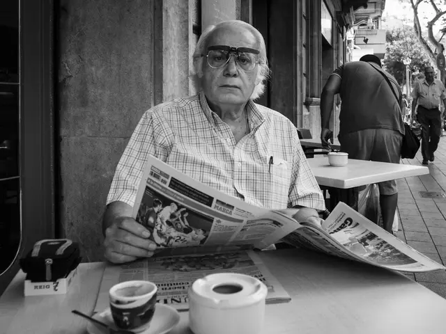 In the image there is an old man sat in front of table with newspaper in hands on a street. back of...