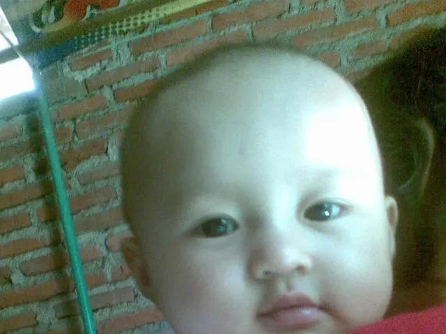 In the picture I can see woman is holding a baby, side there is a brick wall along with one pole.